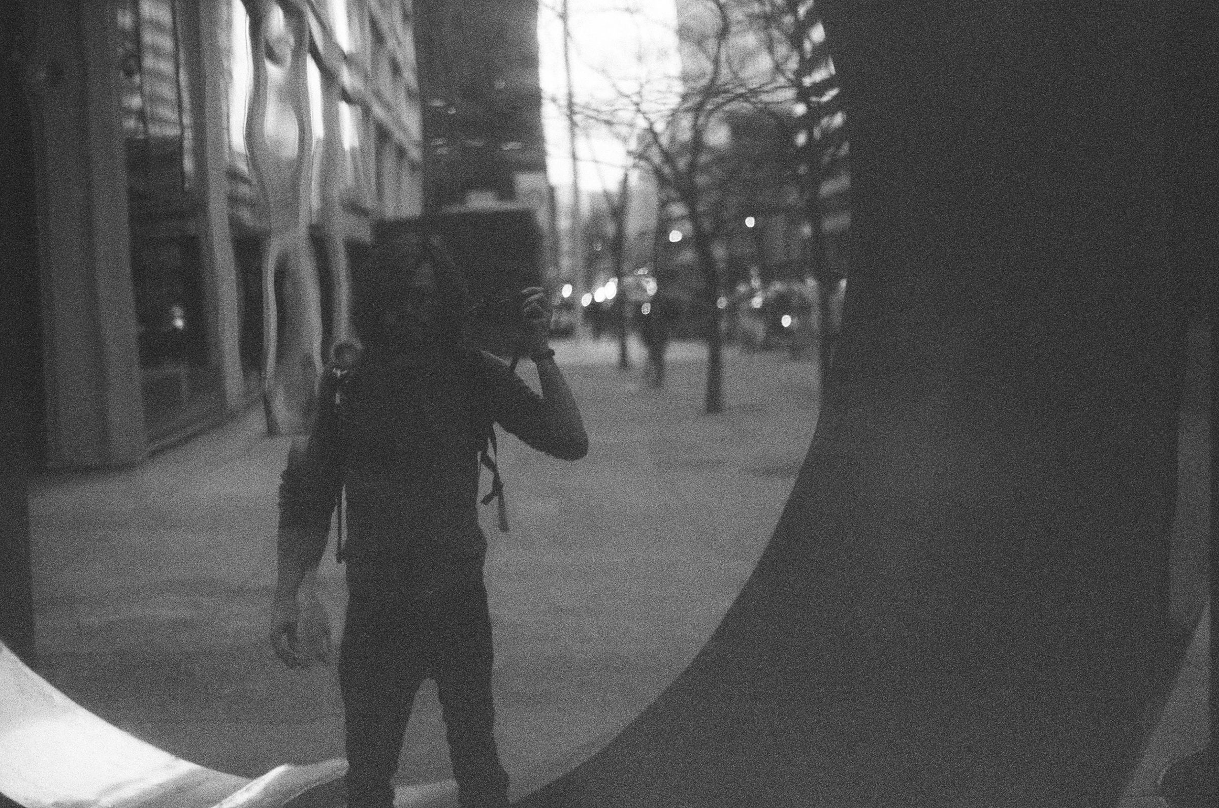 The photographer reflected in a curved mirror on a city street, camera raised